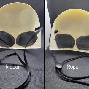 May include: A cream-colored mask with black mesh eye coverings. The mask has a curved shape and is shown with a black ribbon and a black rope. The word "Ribbon" is written on the image.