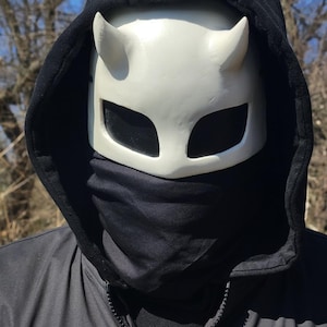 May include: A white horned mask with large eye openings and a black hood. The mask has a smooth surface and is worn with a black neck gaiter and a black hooded jacket.