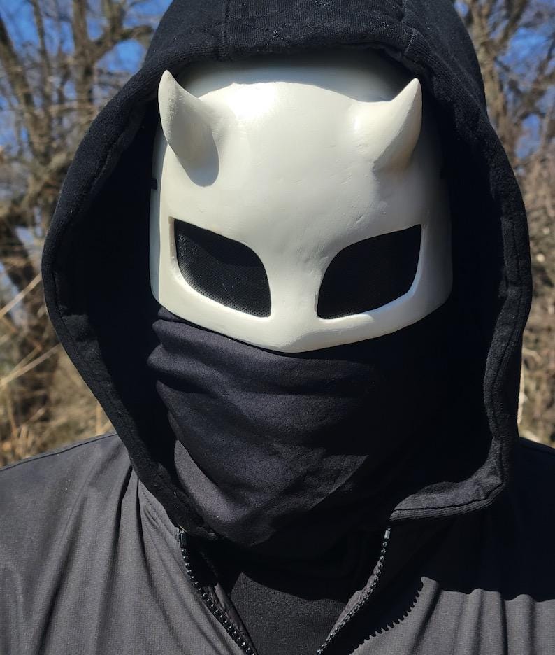 May include: A white horned mask with large eye openings and a black hood. The mask has a smooth surface and is worn with a black neck gaiter and a black hooded jacket.