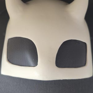 May include: A cream-colored mask with two upward-curving horns and two eye openings covered with black mesh. The mask is made of a smooth, solid material and is designed to cover the upper face.
