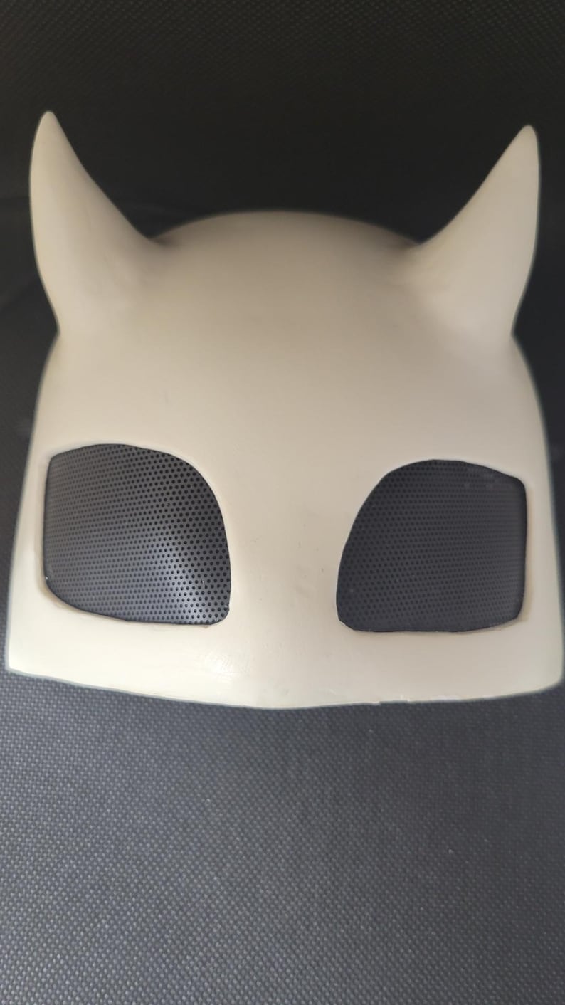 May include: A cream-colored mask with two upward-curving horns and two eye openings covered with black mesh. The mask is made of a smooth, solid material and is designed to cover the upper face.