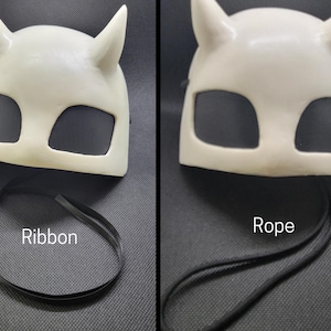 May include: Off-white mask with two upward-curving horns and two eye openings covered with black mesh. The mask has a black ribbon attached. The word "Ribbon" is printed on the image.