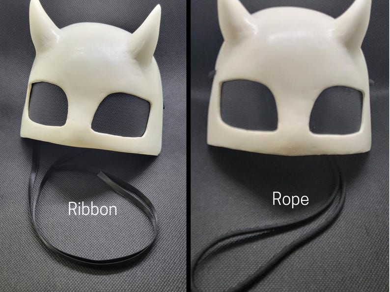 May include: Off-white mask with two upward-curving horns and two eye openings covered with black mesh. The mask has a black ribbon attached. The word "Ribbon" is printed on the image.