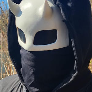 May include: A white horned mask with black mesh eye coverings is worn with a black hooded sweatshirt and a black face covering. The mask has a devil-like appearance. The background is a sunny outdoor scene.