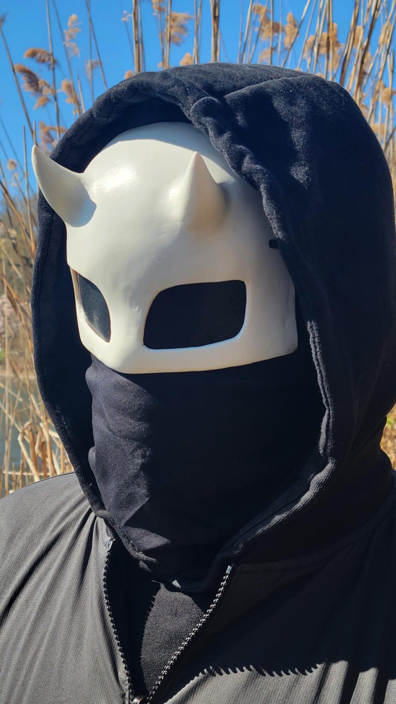 May include: A white horned mask with black mesh eye coverings is worn with a black hooded sweatshirt and a black face covering. The mask has a devil-like appearance. The background is a sunny outdoor scene.