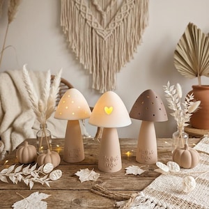 Könnte beinhalten: Drei pilzförmige Lampen mit glatten, matten Oberflächen in Creme-, Hellbeige- und Brauntönen. Die Lampen verfügen über ausgeschnittene Designs, darunter kleine kreisförmige Löcher und eine Herzform, mit eingeprägtem Text am Lampenfuß.
