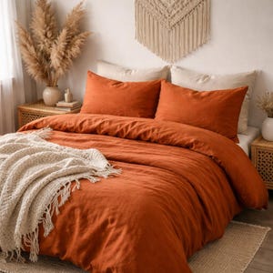 May include: An orange-colored bed set with two matching pillowcases. A cream-colored fringed throw blanket is draped over the bed. The bed is made up with a duvet cover. Decorative items include a vase of dried flowers and a macrame wall hanging.