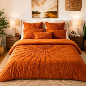 Puede incluir: Un juego de cama color naranja quemado con un patrón de rayos de sol texturizado, formado por líneas con mechones que irradian desde un semicírculo central. Incluye una funda nórdica y cinco almohadas a juego, rectangulares y cuadradas, en color óxido.