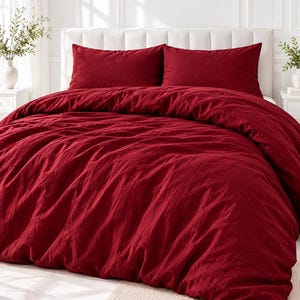 Puede incluir: Un juego de funda nórdica de color rojo intenso con un acabado de tela texturizada. La ropa de cama incluye una funda nórdica grande y arrugada sobre una cama y dos fundas de almohada rectangulares a juego contra un cabecero blanco.