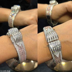 Puede incluir: Una pulsera articulada de tono plateado con un diseño central en forma de lazo incrustado con piedras transparentes de corte baguette. La banda presenta filas de pequeñas piedras redondas transparentes y piedras cuadradas de color rosa claro. W-1139-Pink.