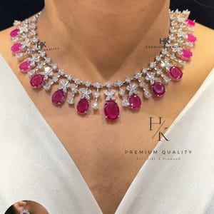 Könnte beinhalten: Eine Halskette mit oval geschliffenen, rosaroten Steinen in silberfarbenem Metall mit klaren Akzentsteinen, dazu passende Ohrhänger. Auf dem Bild ist der Text HK PREMIUM QUALITY sowie N-1109-Ruby zu sehen.