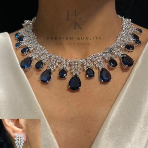 May include: A silver-toned necklace featuring numerous clear marquise-cut stones and multiple teardrop-shaped dark blue stones. The set includes matching drop earrings with a floral-shaped top and a single teardrop-shaped dark blue stone.