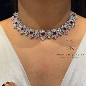 Op de afbeelding: Een sprankelende ketting met bijpassende oorbellen. De ketting heeft vierkante, dieprode edelstenen, geplaatst in een patroon van heldere, briljant geslepen stenen. De oorbellen weerspiegelen het ontwerp van de ketting. De tekst "PREMIUM QUALITY" is zichtbaar.