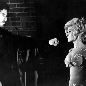 May include: Black and white photograph of a man and a woman. The man, with dark hair and a beard, is reaching towards the woman. The woman is wearing a floral patterned top and has blonde hair. The background features a brick wall.