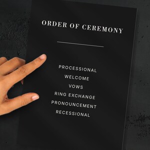 May include: A black wedding ceremony program with white text. The text reads "ORDER OF CEREMONY" at the top, followed by a list of ceremony elements: Processional, Welcome, Vows, Ring Exchange, Pronouncement, and Recessional.