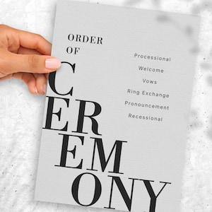 May include: A light grey wedding ceremony program with black text. The words "ORDER OF CEREMONY" are displayed, with a list of ceremony events on the right. The program is held by a hand.