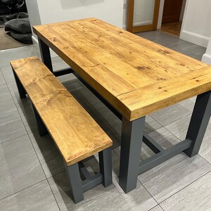 May include: A wooden dining table and bench set. The tabletop and bench seat are made of light brown wood. The table and bench legs are painted a dark grey colour. The table is approximately 183 cm long.