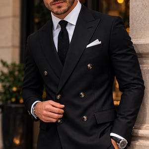 May include: A black double-breasted suit with gold buttons, a white dress shirt, and a black tie. The suit has a pocket square and is worn with a wristwatch. The man is standing outside a building.