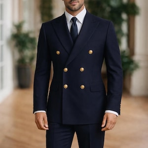 May include: A navy blue double-breasted suit with gold buttons. The suit includes a matching navy tie and a white dress shirt. The man is standing in a well-lit hallway with a wooden floor and green plants in the background.