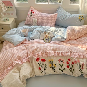 Op de afbeelding: Een bed met een lichtblauwe dekbedovertrek en een roze geruite dekbedovertrek. De dekbedovertrek heeft een witte ruche en geborduurde bloemen. Twee kussens zijn zichtbaar, een roze geruit met rode bloemborduurwerk en een lichtblauw met gele bloemborduurwerk.