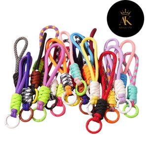May include: A collection of colorful keychains with various rope and cord designs. The keychains feature a variety of colors, including purple, green, orange, and red, with metal rings for attaching keys. The image includes a black circle with the text "AK NEXUS LTD".