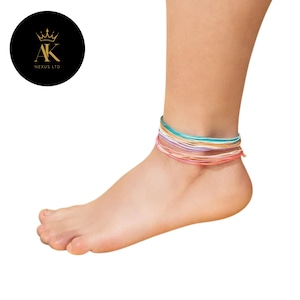 May include: Ankle bracelets in various colors, including turquoise, orange, pink, and purple, are worn on a foot. The bracelets are made of thin, colorful threads. The image is set against a white background.