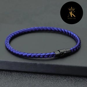 Könnte beinhalten: Ein geflochtenes blaues Armband mit einem schwarzen Verschluss. Das Armband besteht aus einem gewebten Material und ist kreisförmig. Das Bild enthält auch einen schwarzen Kreis mit dem Text "AK NEXUS LTD" und einem Kronenlogo.