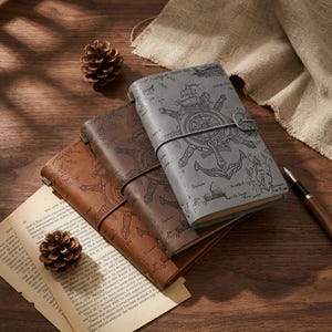 Puede incluir: Tres cuadernos de cuero en tonos marrón y gris, con diseños de temática náutica: anclas, timones y mapas. Los cuadernos se cierran con una correa y una hebilla. También se ven un bolígrafo y una piña.