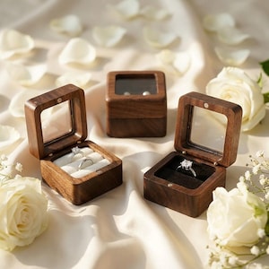 May include: Three square wooden ring boxes with clear lids. Two boxes are open, displaying silver rings with clear stones on white and black velvet inserts. One box is closed. The boxes are arranged among white rose petals and baby's breath.