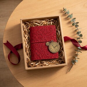 May include: A red journal with a lace overlay texture, secured by a red elastic band featuring a round bronze-toned clock charm. The journal is nestled inside a rectangular cardboard gift box filled with shredded paper filler.