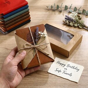 Puede incluir: Un portapasaportes de cuero color canela con un diseño de árbol grabado, envuelto en papel marrón con cordel y lavanda seca. Al lado hay una caja de regalo de cartón marrón con una ventana transparente y una tarjeta que dice Happy Birthday or Safe Travels.