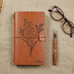 May include: A brown leather journal with a floral design and the name "Alice" engraved at the bottom. A matching wooden fountain pen and eyeglasses are next to the journal on a textured surface.