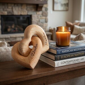 May include: A light-coloured wooden sculpture consisting of two interlocking heart shapes, with the names Dedra and Matthew engraved in a cursive script. The sculpture sits next to a stack of three books and a lit amber glass candle.