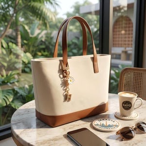 May include: Cream-colored tote bag with brown leather handles and base. A gold charm with a daisy and the letter "H" hangs from the handle. The bag sits on a marble table with a coffee cup, coaster, sunglasses, and a phone.