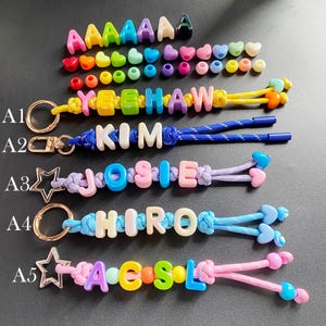 Op de afbeelding: Een verzameling kleurrijke sleutelhangers met kralen die namen en woorden spellen. De sleutelhangers hebben letterkralen, hartvormige kralen en sterrenbedels. De namen zijn "YE HAW", "KIM", "JOSIE", "HIRO" en "ACSL".