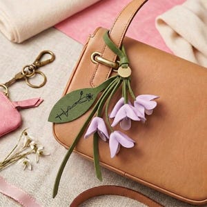 Può includere: Una borsa in pelle marrone chiaro con un ciondolo a forma di fiore in pelle verde. Il ciondolo presenta un'etichetta a forma di foglia con il nome "Amelia" e un motivo floreale, più tre boccioli di fiori color lavanda.