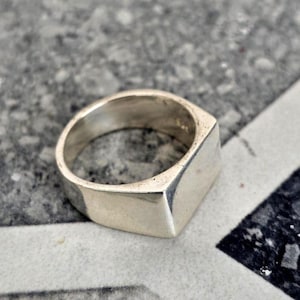 Puede incluir: Un anillo de sello plateado con una cara cuadrada. El anillo está hecho de un material metálico brillante y tiene un diseño simple y moderno. El anillo se muestra sobre una superficie texturizada.