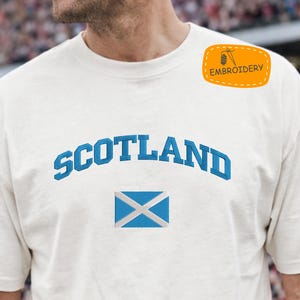 May include: Cream-colored t-shirt with the word "SCOTLAND" arched across the chest in blue embroidery. Below the word is a blue and white embroidered Scottish flag. A small orange patch with the word "EMBROIDERY" is on the upper right.