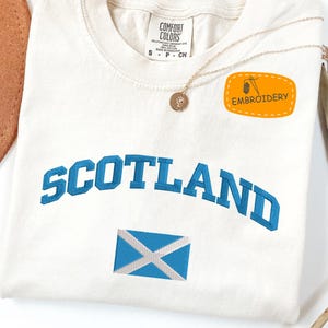 May include: Cream-colored t-shirt with "SCOTLAND" in blue lettering and a blue and white flag emblem. The shirt has a small "EMBROIDERY" patch and a gold necklace with a pendant. The shirt is made by Comfort Colors.