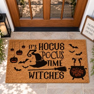 May include: A rectangular tan coir doormat featuring black printed graphics of a witch hat, a broom, hanging jack-o'-lanterns, bats, and a cauldron with legs. The text reads: IT'S HOCUS POCUS TIME WITCHES, with the words Witches Brew on the cauldron.