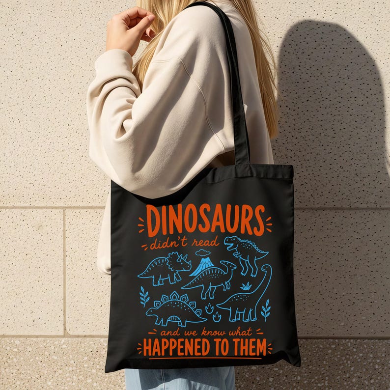 May include: Black tote bag with a blue and orange dinosaur illustration and the text "DINOSAURS didn't read and we know what HAPPENED TO THEM." The bag has a black handle and is made of a durable material.