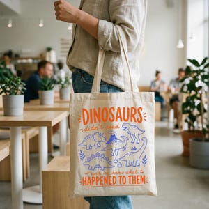 May include: A beige canvas tote bag with orange text that reads "DINOSAURS didn't read and we know what HAPPENED TO THEM." The bag features blue illustrations of various dinosaurs. The bag is being held by a person.