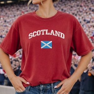 May include: A red t-shirt with the word "SCOTLAND" in white block letters above a small square with a blue and white "X" design. The shirt is short-sleeved and appears to be made of a soft material.