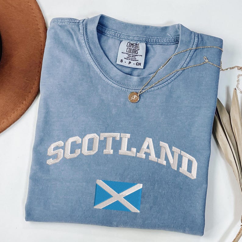 May include: A light blue t-shirt with the word "SCOTLAND" embroidered in white, along with the Scottish flag. A gold necklace with a pendant rests on the shirt. The shirt is made of Comfort Colors material.