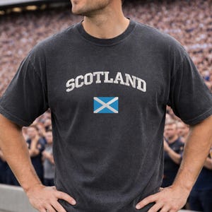 May include: Dark gray t-shirt with the word "SCOTLAND" in white arched lettering above a blue and white flag. The shirt has a crew neck and short sleeves.