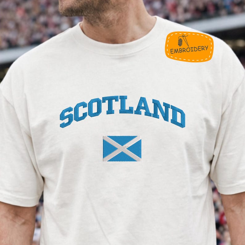 May include: Cream-colored t-shirt with the word "SCOTLAND" arched across the chest in blue embroidery. Below the word is a blue and white embroidered Scottish flag. A small orange patch with the word "EMBROIDERY" is on the upper right.