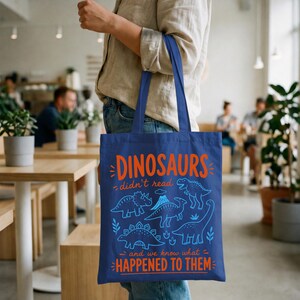 May include: A navy blue tote bag featuring orange and blue text and dinosaur illustrations. The text reads "DINOSAURS didn't read and we know what HAPPENED TO THEM". The bag has two navy blue handles.