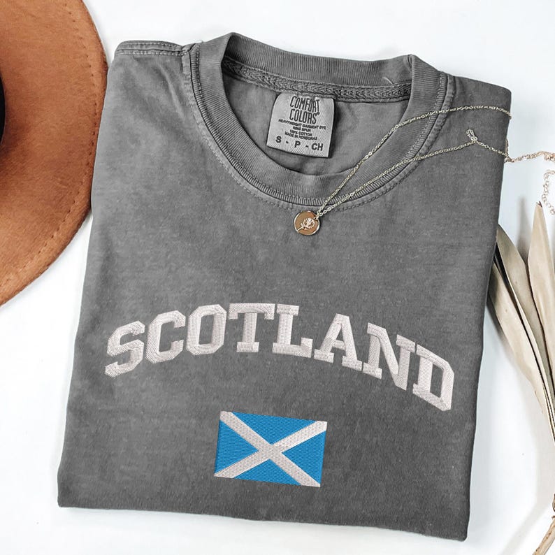 May include: A gray Comfort Colors t-shirt with the word "SCOTLAND" embroidered in white, and a blue and white flag emblem. A silver necklace with a pendant is draped across the shirt. The shirt is folded on a white surface.