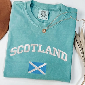 May include: A turquoise Comfort Colors t-shirt with the word "SCOTLAND" embroidered in white, along with the Scottish flag. A gold necklace with a pendant rests on the shirt. The shirt is size small.