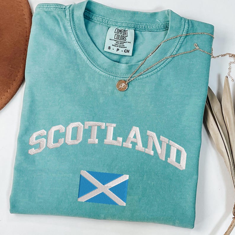 May include: A turquoise Comfort Colors t-shirt with the word "SCOTLAND" embroidered in white, along with the Scottish flag. A gold necklace with a pendant rests on the shirt. The shirt is size small.